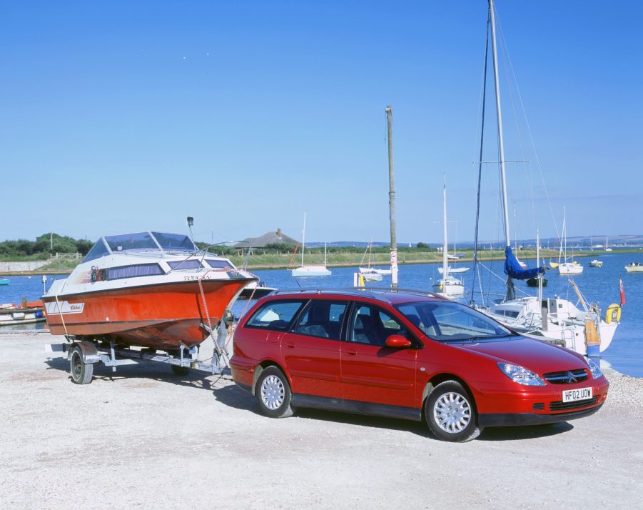 A red 2002 Citroen C5 hdi crossover towing a boat by a harbor