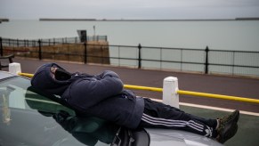 A man wearing a facemask sleeps on the hood of his car, overlooking a large body of water