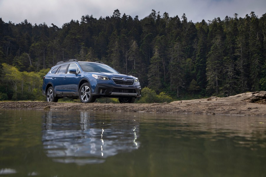 A blue 2020 Subaru Outback midsize SUV parked by a lake and pine trees