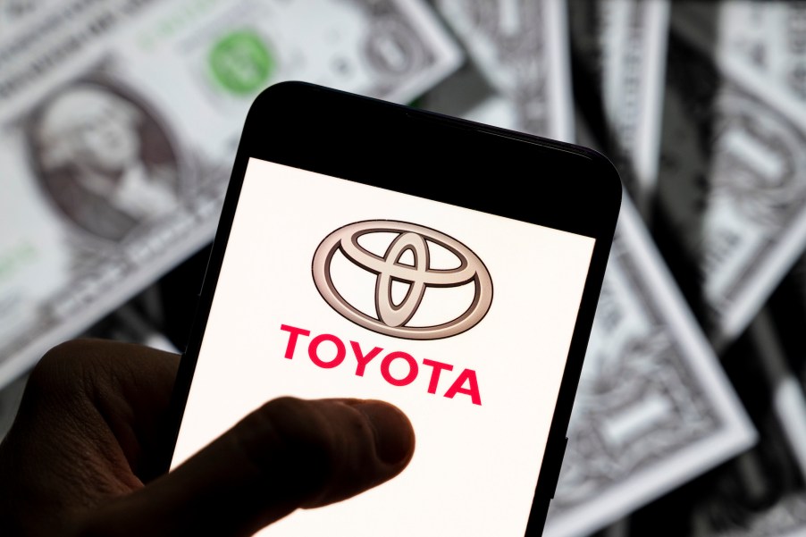 A photo illustration of the Toyota logo displayed on a smartphone screen with $1 bills in the background