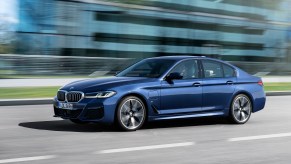 A blue metallic 2021 BMW 530e hybrid sedan traveling past a glass building on a city street on a sunny day