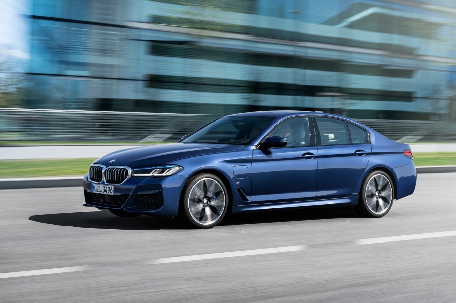 A blue metallic 2021 BMW 530e hybrid sedan traveling past a glass building on a city street on a sunny day