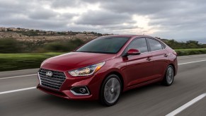 A red metallic 2021 Hyundai Accent traveling on a highway past green grass and hills on a cloudy day