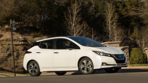 A white 2021 Nissan Nissan Leaf EV parked on a hilly suburban street with trees and a rock wall behind it