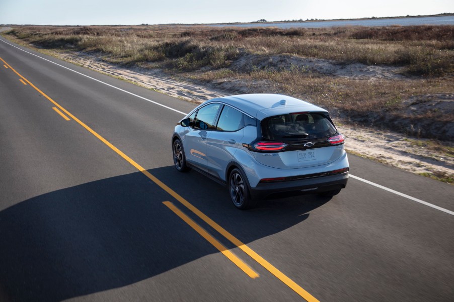 A silver 2022 Chevy Bolt EV travels on a two-lane highway along a large body of water on a sunny day