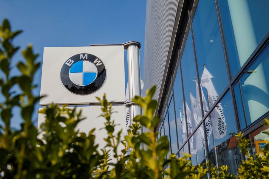 A BMW dealership marque is beside of a building with large windows with some blurred green shrubbery in the front.