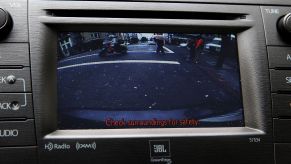 A infotainment screen with a backup camera view that has pedestrians and motorcyclists in the view of the camera with 'Check surroundings for safety' in red at the bottom of the screen.