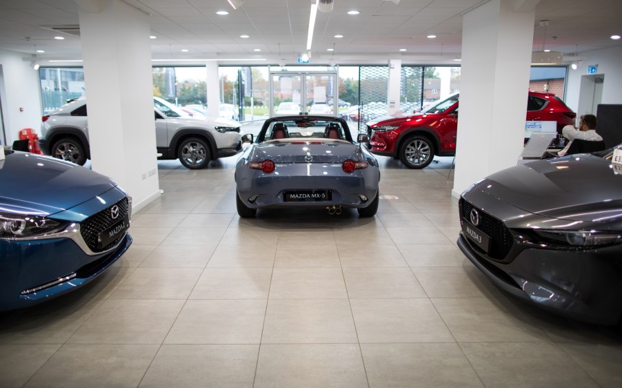 Various Mazda cars sit in a dealership style room with the back end of a grey Mazda MX-5 in the center, the front hood of a Mazda 2 on the right and a Mazda 3 on the left, with two crossover Mazda's in the back partially blocked by white pillars. All in front of a wall length window.