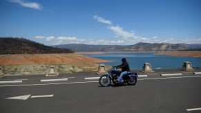 Motorcyclist riding on a black bike dressed in black over a bridge in California with a Mountainous landscape in the background and a brilliant blue body of water.