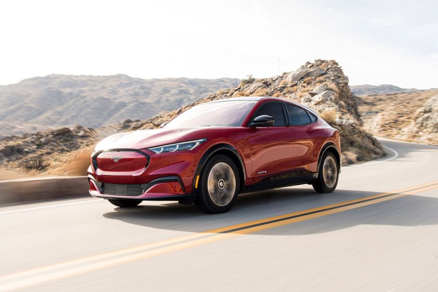 A red 2021 Ford Mustang Mach-E is driving on a highway with a brown rocky scene in the background.