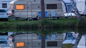 A cream and gray travel trailer with a single tree in the middle front, parked beyond a pond where the reflection of the RV can be seen. There are two darker blue and one lighter blue lawn chair in front of the travel trailer. There are other cars and travel trailers in the background.