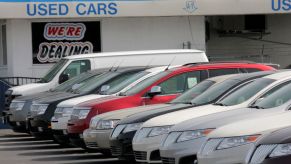 A picture of a used car lot similar to a buy here pay here with numerous cars of various colors in front of a small white building with a blue overhang that says "used cars" with a sticker in a blacked out window that says "We're Dealing."