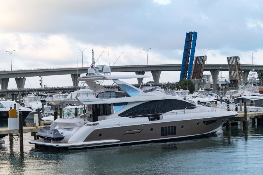 A smaller white and grey yacht parked in a marina with numerous yachts and a bridge in the background