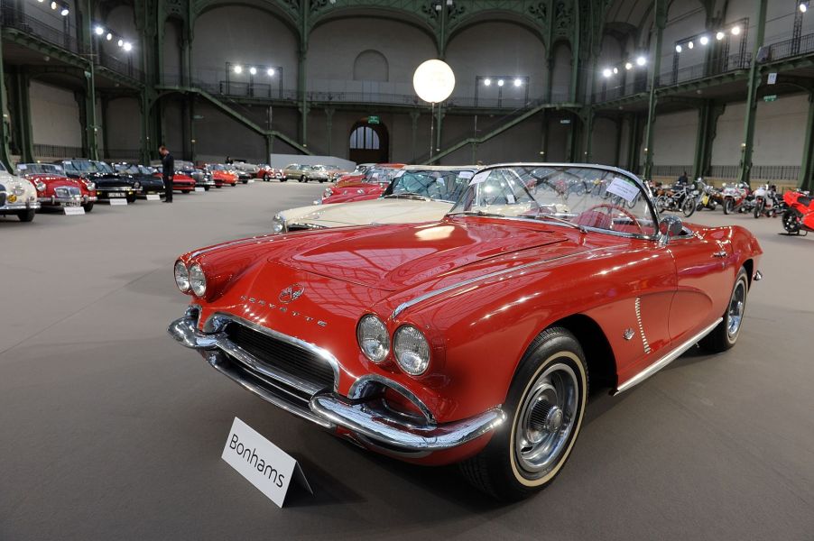 A red 1962 Chevrolet Corvette sitting in a large building with high ceilings with other classic cars.