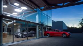 A red 2020 Chevrolet Malibu sits outside at twilight in front of a building with glass windows with a grey 2020 Chevrolet Malibu sitting behind the red one inside.