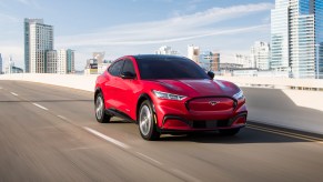 A red 2021 Ford Mustang Mach-E electric crossover SUV traveling on a bridge overlooking a city skyline