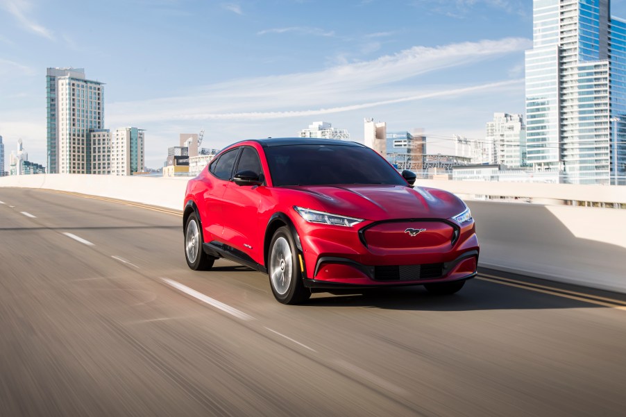 A red 2021 Ford Mustang Mach-E electric crossover SUV traveling on a bridge overlooking a city skyline