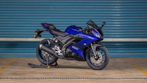 A blue-and-black 2021 Yamaha YZF-R15 on a rear-wheel stand by a turquoise garage