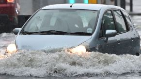 A blue hatchback driving through flood waters that come almost over the headlights.