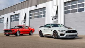 A white 2021 Mustang Mach 1 sitting in front of an older orange model in front of a metal garage style building.