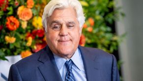 Jay Leno wearing a blue suit with a light blue shirt and a dark stripped blue tie in front of a flowered background.