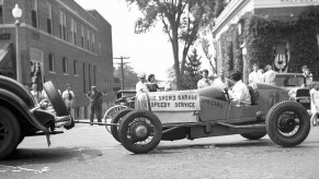 A black-and-white photo of an early racecar being towed by a rope through a town