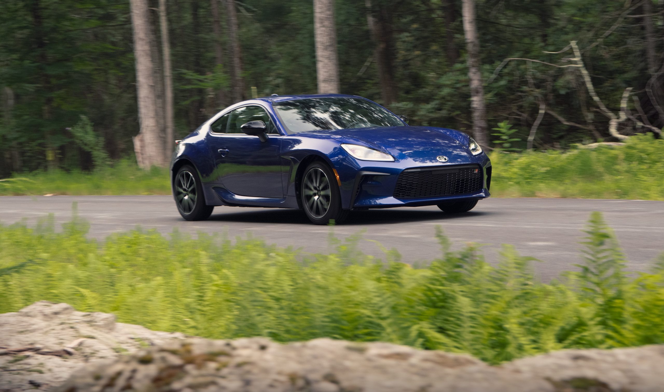 A dark blue 2022 Toyota GR 86 rolls down a forest road, shot from the front 3/4