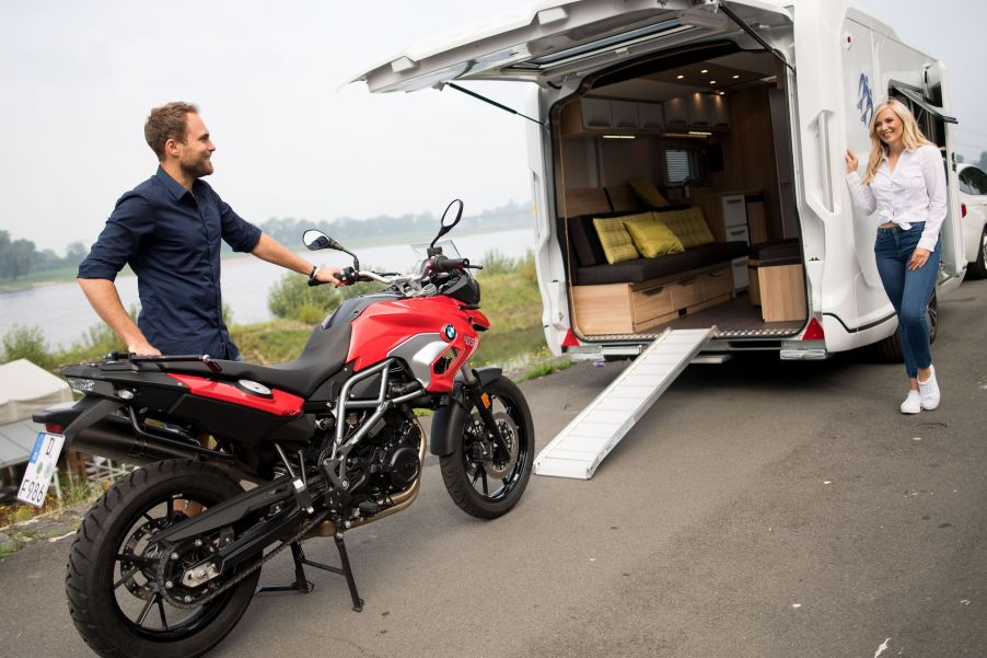 A man loads a red BMW motorcycle onto a white Knaus camper van RV with a ramp