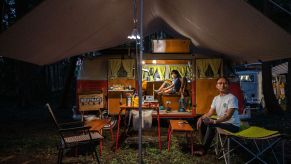 A set up camper van with a canopy outside, a young child sitting inside and a man outside in a chair.