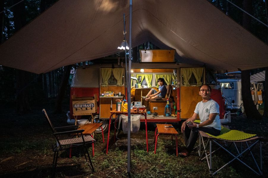 A set up camper van with a canopy outside, a young child sitting inside and a man outside in a chair.