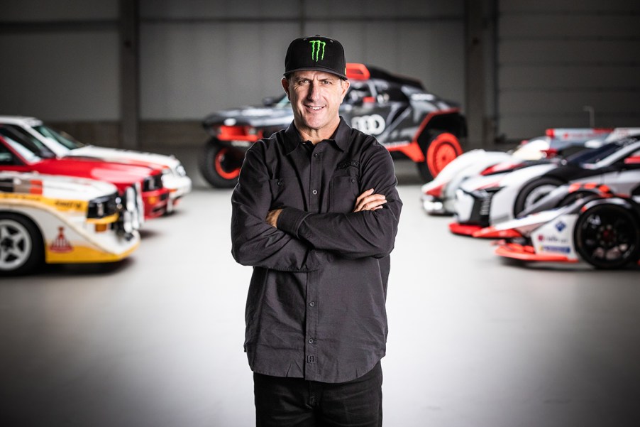 Rally racer Ken Block standing in a room with Audi rally cars and other race cars behind him