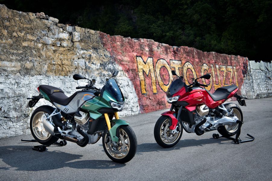 A green-and-silver and a red Moto Guzzi V100 Mandello on rear-wheel stands by a graffiti-covered brick wall