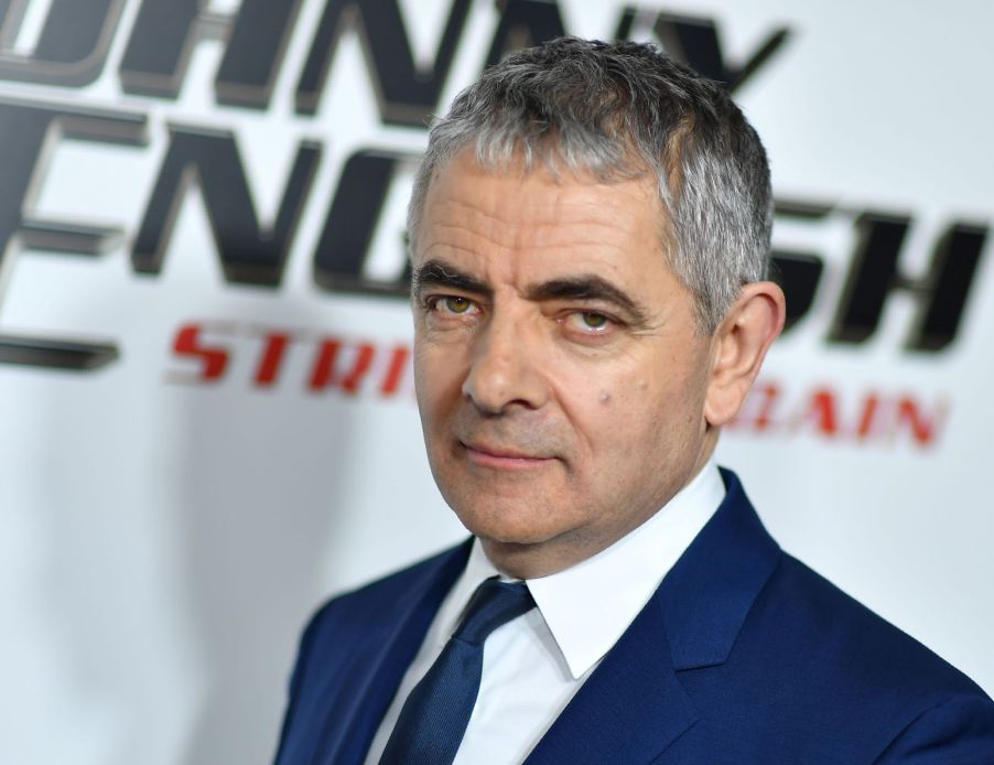 Rowan Atkinson dressed in a blue suit standing in front of a white background with black and red writing.