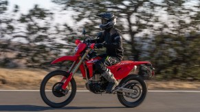 A black-clad rider takes a red 2022 Honda CRF450L dirt bike down a paved road