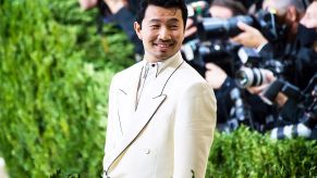 Simu Liu in a white suite with photographers in the background surrounded by shrubbery.