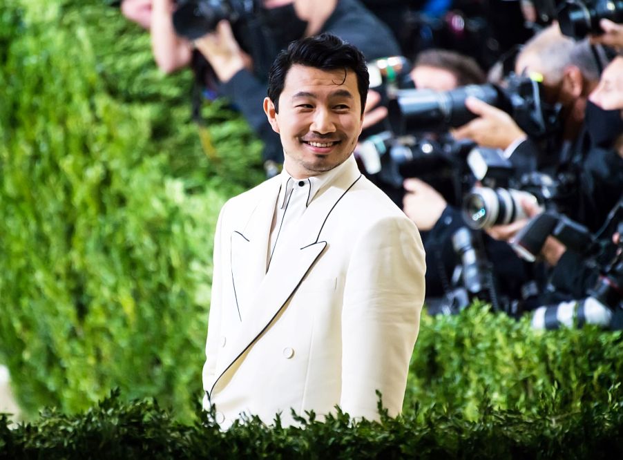 Simu Liu in a white suite with photographers in the background surrounded by shrubbery.