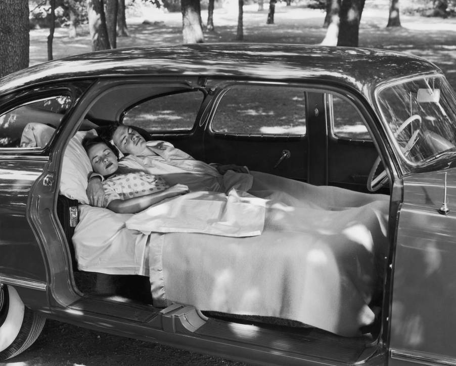 A couple sleeps in an antique car with fold-flat seats in a residential neighborhood.