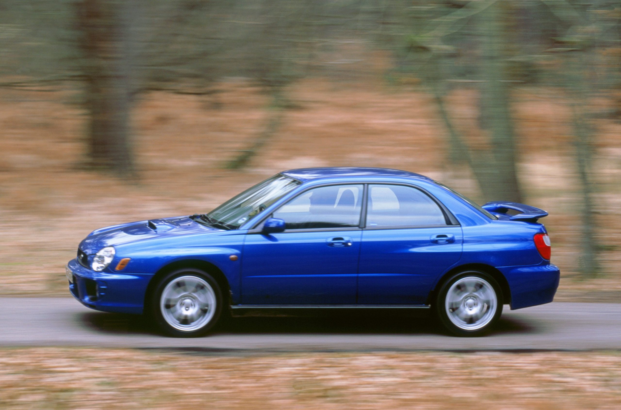 A blue Impreza WRX shoots down a country road, shot in profile.