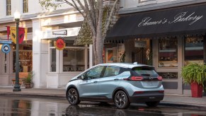 A light-blue metallic 2022 Chevrolet Bolt EV parked on a wet city street outside a bakery