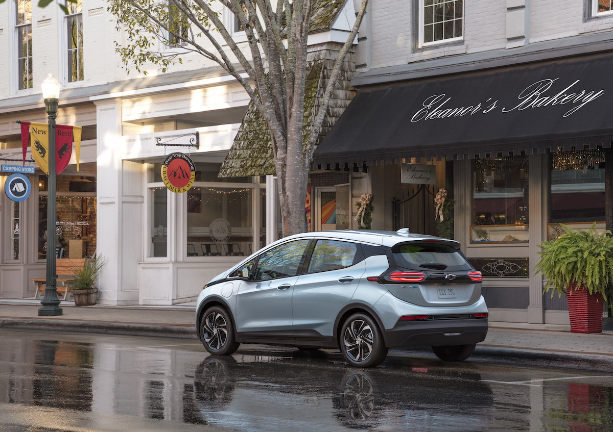 A light-blue metallic 2022 Chevrolet Bolt EV parked on a wet city street outside a bakery