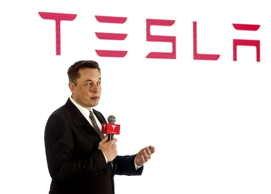 Tesla, creator of autopilot, speaking in front of a white background with Tesla written in red at the top dressed in a suit.