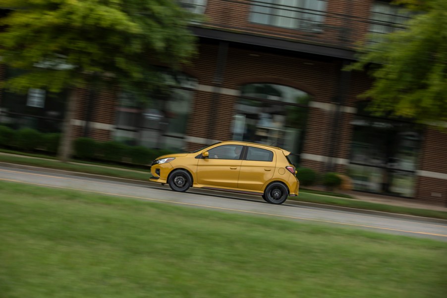 A yellow 2021 Mitsubishi Mirage subcompact hatchback traveling past a red-brick building, trees, and grass