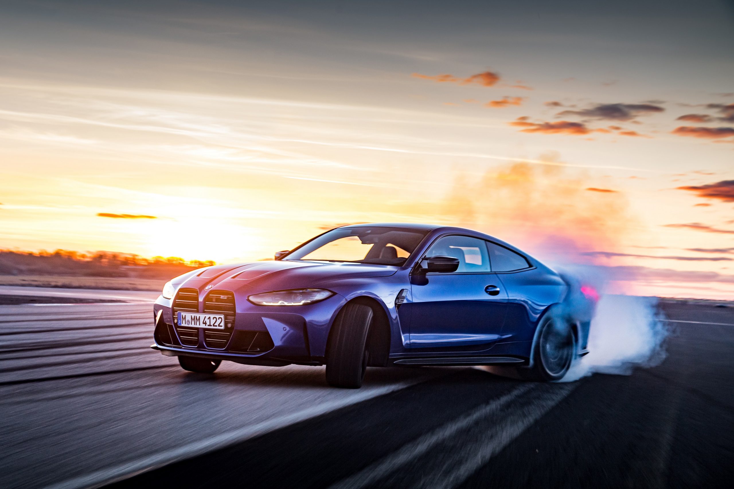 A blue 2022 BMW M4 drifts through a turn at sunset