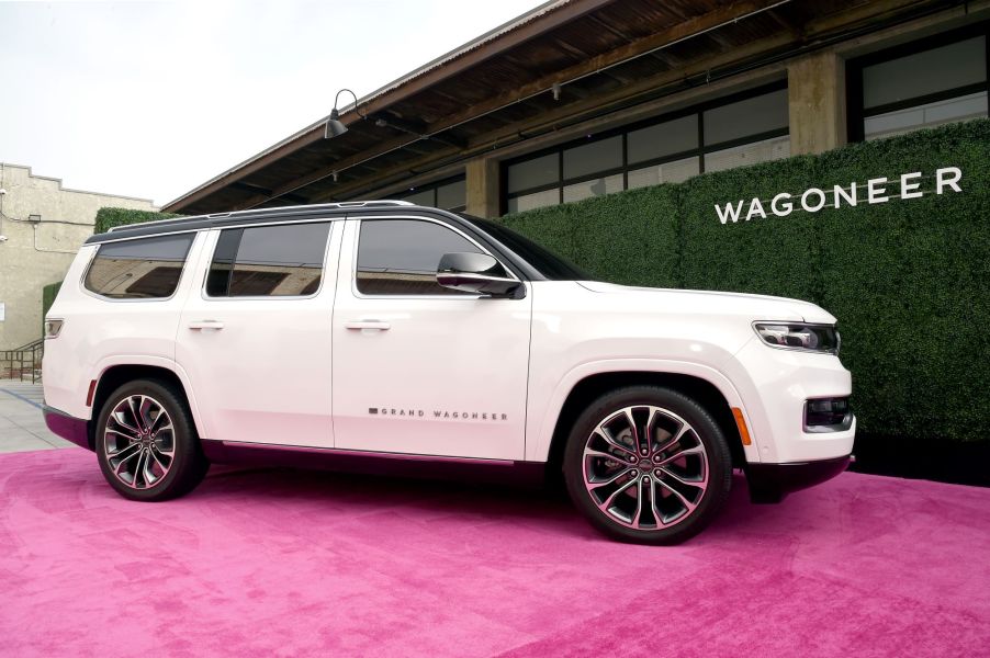 A white white Jeep Grand Wagoneer, maker of the Jeep Wagoneer trim, on a light red carpet in front of a black and brown building with Wagoneer printed in white on the side the building.