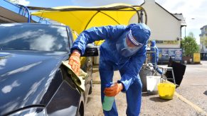 A person washing a car as part of a black car wash dressed in a blue hazmat style suit in an outdoor setting with a yellow tent in the background.