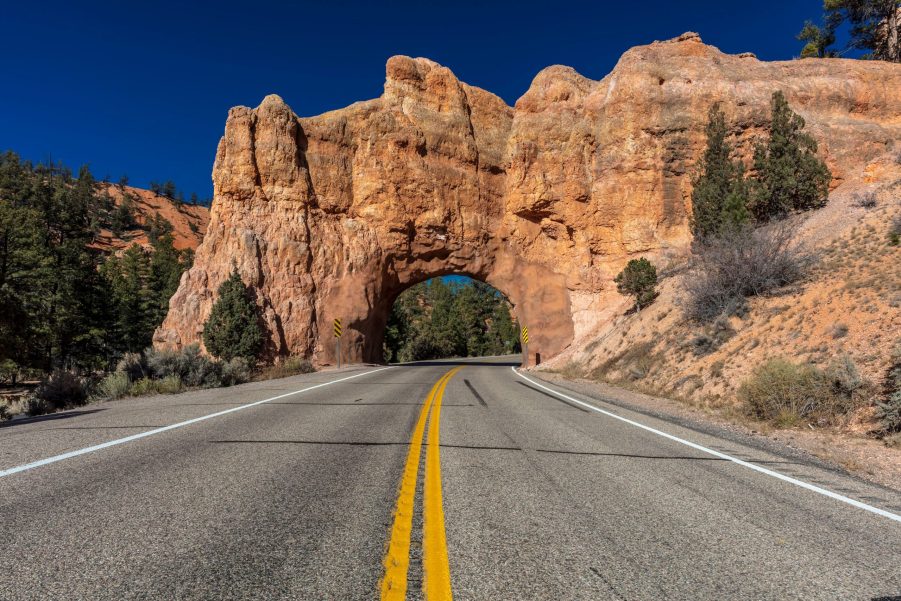 A road with a double solid yellow line in a rocky area with a tunnel ahead made of rocks.