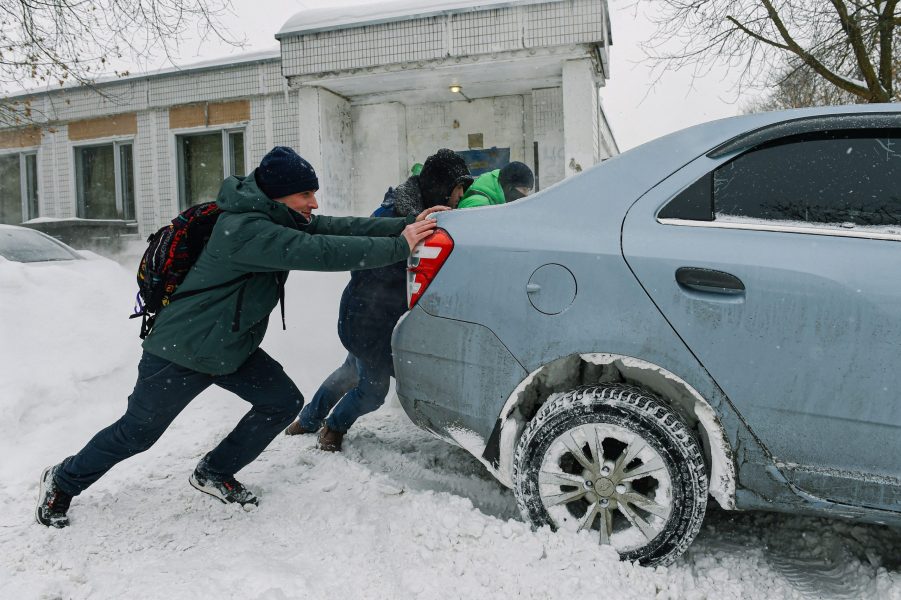 A group of people pushing a light blue car stuck in the snow.