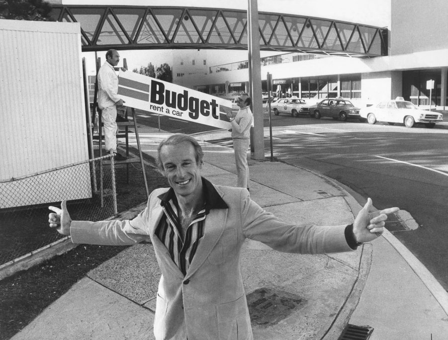 man standing in front of a sign reading "Budget car rentals" although the cost of a rental car has skyrocketed over the last few years.