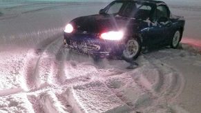 A Mazda Miata roadster parked in a circle of tire tracks left in a snow-covered parking lot.