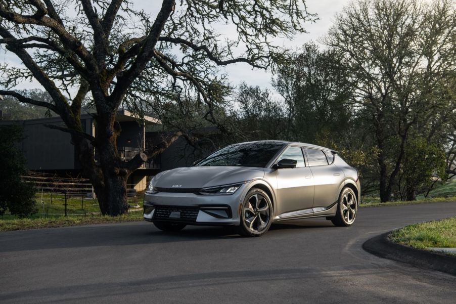 A silver 2022 Kia EV6 surrounded by trees with a building in the background on a blacktop road.